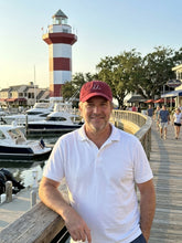 Man in a red cap and white polo shirt standing on a dock with a lighthouse in the background.