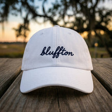 White cap with 'bluffton' text on a wooden surface with a blurred natural background