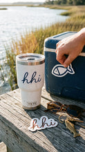 Hand applying a fish sticker to a navy bag beside an hhi tumbler and stickers on a marsh-side rail.