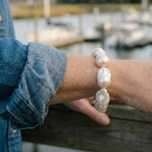 A close-up of a person's wrist wearing a bracelet made of large, irregular white baroque pearls, with a blue denim shirt rolled up and a blurred outdoor waterfront background.