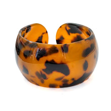 A wide, chunky cuff bracelet made of glossy tortoiseshell resin, featuring a pattern of amber orange and black, set against a plain white background.