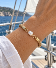 A gold-plated chain bracelet with a central white baroque pearl and gold spacer rings, worn on a person's wrist on a sunny day at a sailboat with the sea in the background.