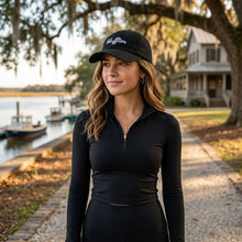 Woman wearing a black cap and long-sleeve top standing by a lake with boats and a house in the background.