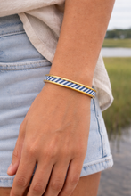 Person wearing a blue and gold striped bracelet by a body of water.