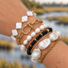 A close-up of a person's wrist wearing a stack of bohemian-style bracelets, including baroque pearls, black beads, a gold chain, and a gold rope bracelet, with a blurred marsh landscape in the background.