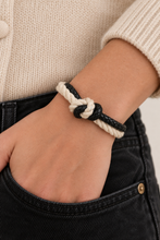 A close-up of a person's wrist wearing a braided rope bracelet with a knot detail, styled with a cream knit sweater and black denim jeans.