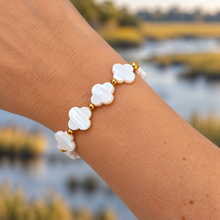 A close-up of a person's wrist wearing a bracelet made of white mother-of-pearl four-leaf clover beads separated by small gold spacer beads, with a blurred outdoor water landscape in the background.