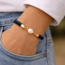 A close-up of a person's wrist wearing a dark navy braided cord bracelet with gold-toned metal caps and a single white pearl bead.