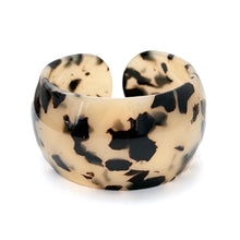 A thick, chunky open cuff bracelet made of glossy, marbled resin with a beige base and black organic splatters, shown on a plain white background.