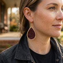 A woman wearing a teardrop-shaped dark red textured stone earring with a gold border and gold hook, shown in a close-up profile shot outdoors. GAMECOCKS