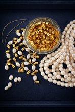 A clear glass jar filled with gold cylindrical bead caps, surrounded by scattered white pearl beads and several strands on a dark navy background.