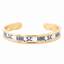 A gold-plated cuff bracelet featuring a white inlay printed with the navy blue text 'HHI, SC' repeatedly, shown on a plain white background.