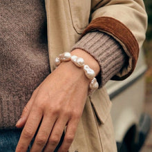 A close-up of a person's wrist wearing a bracelet made of irregular white baroque pearls, styled with a beige jacket and brown ribbed sweater.