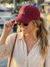 Woman wearing a red cap with 'hii' text, sunglasses, and a beige shirt by a waterfront.