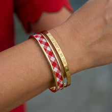 Close-up of a wrist wearing a red and white checkered bracelet and gold band with engraved text.