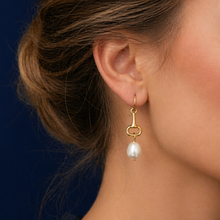 Gold earring with a pearl drop worn by a person against a dark blue background