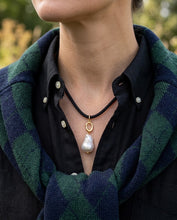 A close-up of a person wearing a black cord necklace with a gold ring pendant and a large white baroque pearl. The person is also wearing a dark shirt and a green and navy plaid scarf, with a blurred outdoor background.