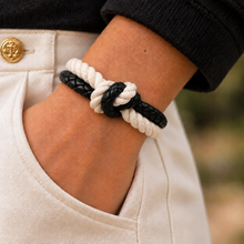 A close-up of a person's wrist wearing a double-strand rope knot bracelet with one white rope and one black braided rope, styled with a black long-sleeve top and off-white trousers.