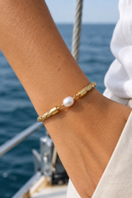 A gold-toned bracelet with a single white pearl bead, worn on a person's wrist on a boat with the blue ocean in the background.