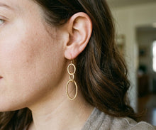A close-up side profile of a woman wearing a pair of gold dangle earrings with one smooth oval hoop and one textured oval drop, shown in a softly blurred indoor setting.