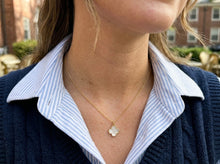 A close-up lifestyle shot of a woman wearing a delicate gold chain necklace with a small white clover pendant, styled over a blue and white striped collared shirt and a navy blue sweater.