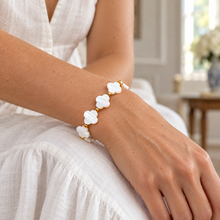 A close-up of a woman's wrist wearing a delicate bracelet made of white mother-of-pearl clover-shaped links with gold-tone metal connectors, set against a soft, bright indoor background.