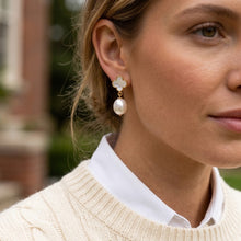 A close-up of a woman wearing a gold and pearl drop earring with a clover-shaped top, shown in an outdoor setting.