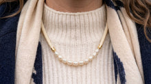 A close-up of a person wearing a pearl necklace with gold-toned spacer beads over a cream ribbed sweater and a cream-and-navy patterned scarf.