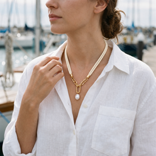 A woman wearing a gold chain necklace with a white pearl pendant and a cream cord detail, styled with a white linen shirt against a blurred marina background.