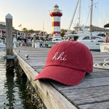 Red cap with 'hhi' on a dock with harbour town lighthouse and boats in the background of sea pines
