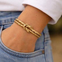 A close-up of a person's wrist wearing a gold braided knot bracelet, with a white shirt and light blue jeans visible.