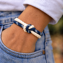 A close-up of a person's wrist wearing a double-strand rope bracelet with a knotted center. The bracelet features one cream-colored rope and one navy blue rope with white woven accents.