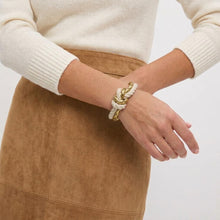 A close-up of a person wearing a cream and gold knotted rope bracelet on their wrist, styled with a cream ribbed sweater and a brown suede skirt against a light gray background.