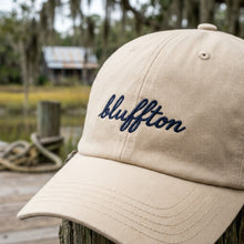 Beige cap with 'bluffton' embroidery on a wooden post by a body of water.