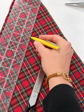 A close-up of a hand holding a yellow pen and measuring tape over a red tartan plaid fabric, with scissors visible on a white surface.