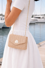 Person wearing a white dress holding a straw handbag with a decorative element by a waterfront.