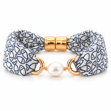 A fabric bracelet featuring a white base with a navy blue fish pattern, accented by polished gold-tone metal rings, a central white pearl, and two gold cylindrical beads at the clasp.