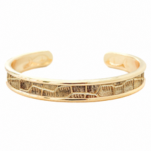 A gold-toned open cuff bracelet featuring a textured, geometric inlay pattern along the front edge, set against a plain white background.
