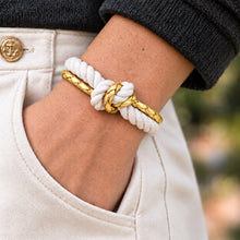 A close-up of a person's wrist wearing a white rope bracelet with a gold braided accent, tucked into a light beige pocket. The person is wearing a dark gray knit sweater and light beige trousers.