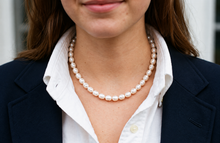 A close-up of a woman wearing a classic white freshwater pearl necklace, styled with a white collared shirt and a dark navy blazer.