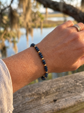 A close-up of a person's wrist wearing a beaded bracelet made of dark blue beads separated by small gold spacer beads, photographed outdoors near a body of water with a wooden railing in the background.