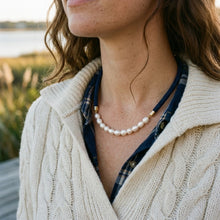 A close-up lifestyle shot of a woman wearing a pearl necklace with gold-toned spacer beads over a navy blue plaid shirt and a cream cable-knit sweater, with a soft-focus outdoor background.