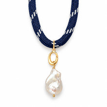 A close-up of a necklace featuring a thick navy blue braided cord with white woven stripes, a gold ring connector, and a large, irregular white baroque pearl pendant on a plain white background.