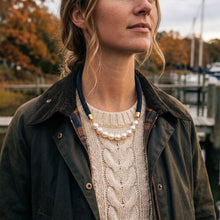 A woman wearing a layered necklace featuring a strand of white pearls and a dark blue beaded cord with gold-toned metal accents, styled over a cream cable-knit sweater and an olive green jacket, set against a soft-focus outdoor background.