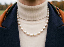 A close-up of a person wearing a double-strand pearl necklace with small gold spacer beads over a cream ribbed turtleneck sweater, a navy coat, and a patterned scarf.