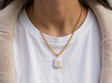 A close-up of a person wearing a delicate gold beaded chain necklace with a small white shell pendant, styled over a white t-shirt and beige cardigan.