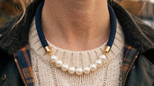 A close-up of a person wearing a statement necklace with white faux pearls and a navy blue braided cord, styled over a cream knit sweater and a dark jacket.