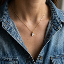 A close-up of a person wearing a delicate gold chain necklace with a small clear rectangular pendant, styled with a blue denim shirt.