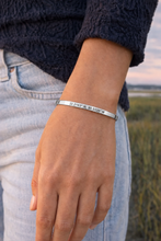 A silver metal cuff bracelet engraved with geographic coordinates, worn on a person's wrist. The person is wearing a dark navy textured sweater and light-wash blue jeans, with an outdoor field in the background. Hilton head isalnd