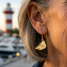 Close-up of a person wearing a gold earring with a 'Klaipéda' charm, blurred lighthouse and boats in the background.